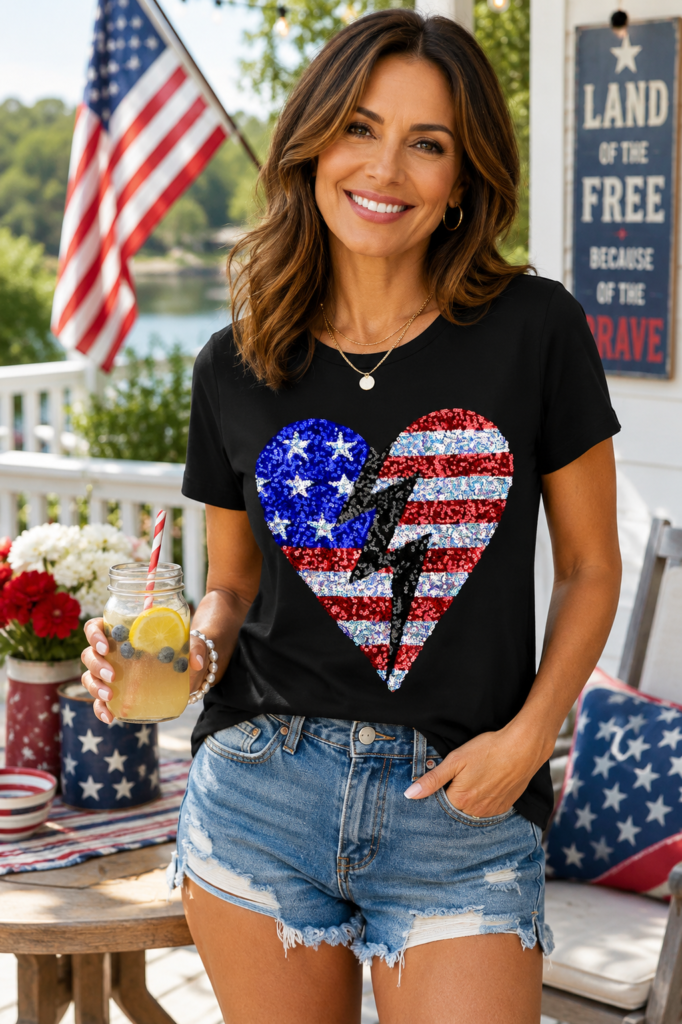 Lifestyle image of a woman wearing a black patriotic sequin heart American flag T-shirt with denim shorts while enjoying a festive outdoor Fourth of July gathering decorated with American flags and summer holiday décor.
